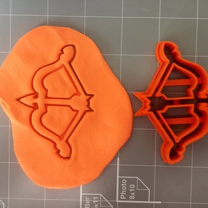 May include: A red plastic cookie cutter in the shape of an arrow and bow. The cutter is shown on a gray surface with a piece of orange modeling clay.