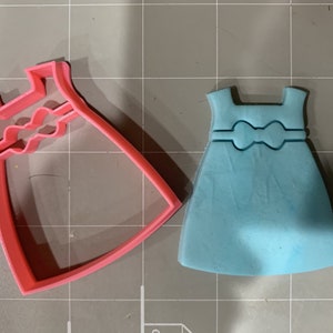 May include: A pink plastic cookie cutter shaped like a dress with a bow. Next to it is a light blue fondant cookie shaped like a dress with a bow.