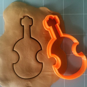 Pode incluir: Cortador de biscoitos de plástico laranja em forma de violino. O cortador está em cima de uma peça de massa de modelar marrom.
