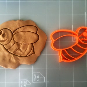 May include: Orange plastic bee-shaped cookie cutter. The cutter is shown on a gray grid surface with a brown clay impression of the bee.