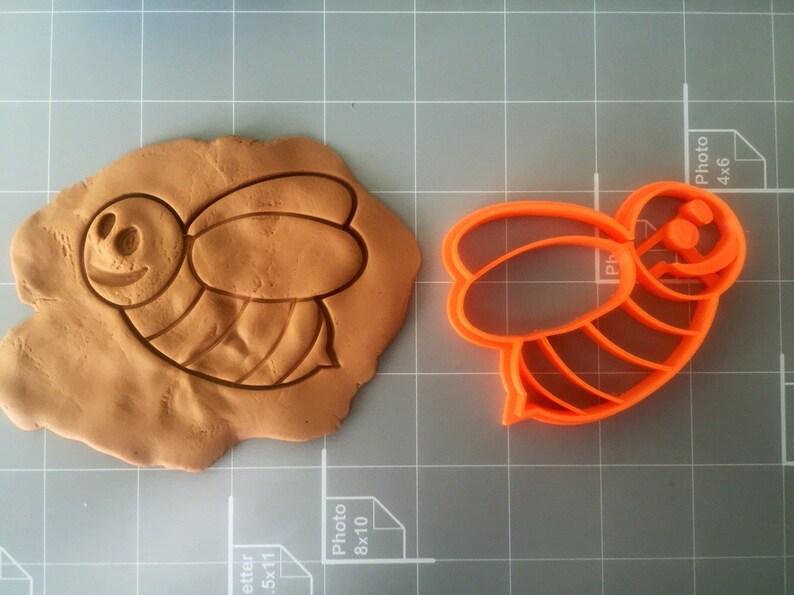 May include: Orange plastic bee-shaped cookie cutter. The cutter is shown on a gray grid surface with a brown clay impression of the bee.