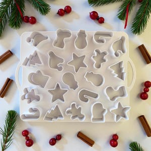 May include: A white plastic cookie cutter tray with various Christmas-themed shapes, including stars, trees, and mittens. The tray is surrounded by cinnamon sticks, red berries, and evergreen sprigs, creating a festive holiday scene.