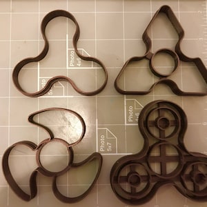 May include: Four brown plastic cookie cutters shaped like fidget spinners. The cutters are on a grey grid surface.