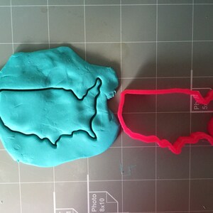 May include: A pink plastic cookie cutter in the shape of the United States of America. The cookie cutter is on a gray surface with a blue piece of modeling clay that has been cut out with the cookie cutter.