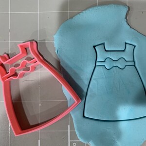 May include: A pink plastic cookie cutter shaped like a dress with a bow. The cutter is on top of blue fondant, which has been cut out in the shape of the dress.
