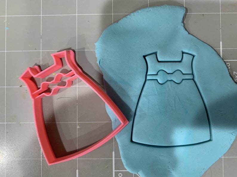 May include: A pink plastic cookie cutter shaped like a dress with a bow. The cutter is on top of blue fondant, which has been cut out in the shape of the dress.
