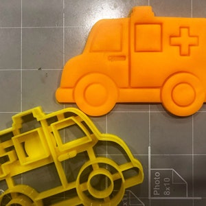 May include: Two yellow plastic cookie cutters shaped like ambulances. The top cutter is orange and has a red cross on the side.