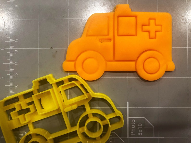 May include: Two yellow plastic cookie cutters shaped like ambulances. The top cutter is orange and has a red cross on the side.
