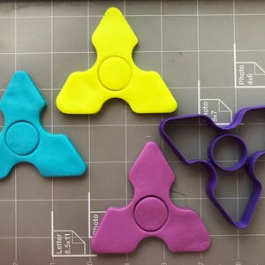 May include: Three different coloured plastic fidget spinners, one yellow, one blue, and one purple, and a purple plastic fidget spinner cookie cutter.