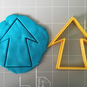 May include: A blue clay impression of an upward pointing arrow next to a yellow plastic cookie cutter in the shape of an arrow.