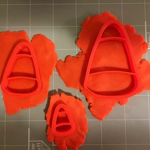 May include: Three red candy corn cookie cutters of different sizes on a gray grid surface.