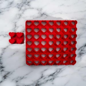 May include: A red silicone mold with multiple heart-shaped cutouts, alongside a smaller version with four hearts. The mold is designed for creating heart-shaped items, perfect for baking or crafting. The background is a white marble surface.
