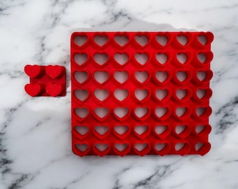 May include: A red silicone mould featuring numerous heart-shaped cutouts, with a smaller version holding four hearts. This mould is designed for creating heart-shaped items, suitable for baking or crafts. The background is a white marble surface.