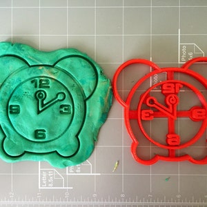 May include: A red plastic cookie cutter shaped like an alarm clock. The cookie cutter is on a gray grid surface.