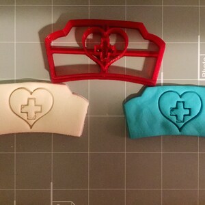 May include: A red plastic cookie cutter in the shape of a nurse's hat with a heart and cross in the center. Two cookies are shown, one white and one blue, with the same design.
