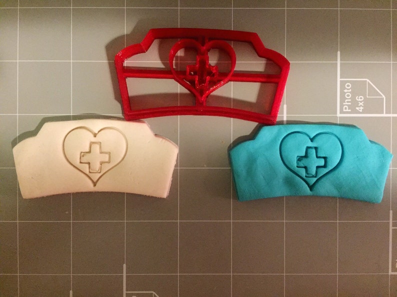 May include: A red plastic cookie cutter in the shape of a nurse's hat with a heart and cross in the center. Two cookies are shown, one white and one blue, with the same design.