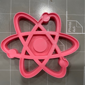 May include: A pink plastic cookie cutter shaped like an atom. The atom has a hexagon in the center and three orbiting electrons.