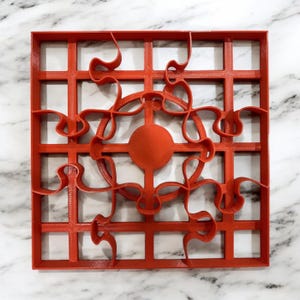 Puede incluir: Un cortador de galletas de plástico rojo con un patrón de cuadrícula y un diseño decorativo en el centro. El diseño presenta un círculo con una serie de bucles y curvas.