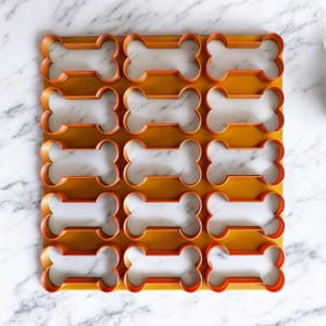 Puede incluir: Un juego de 15 cortadores de galletas con forma de hueso de color naranja sobre una superficie de mármol blanco.
