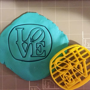 K&ouml;nnte beinhalten: Ein gelber Plastik-Ausstecher in Form des Wortes "LOVE" mit einem Gittermuster. Ein St&uuml;ck t&uuml;rkisfarbenes Modellierton befindet sich hinter dem Ausstecher.