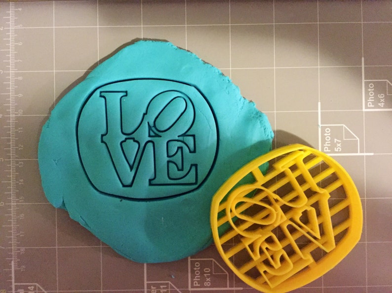 K&ouml;nnte beinhalten: Ein gelber Plastik-Ausstecher in Form des Wortes "LOVE" mit einem Gittermuster. Ein St&uuml;ck t&uuml;rkisfarbenes Modellierton befindet sich hinter dem Ausstecher.