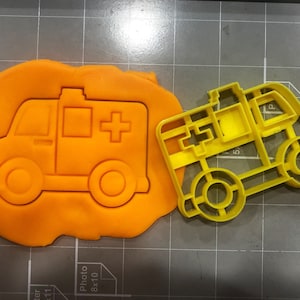 May include: Yellow plastic cookie cutter shaped like an ambulance. The cutter has a red cross on the side and a white rectangular window.