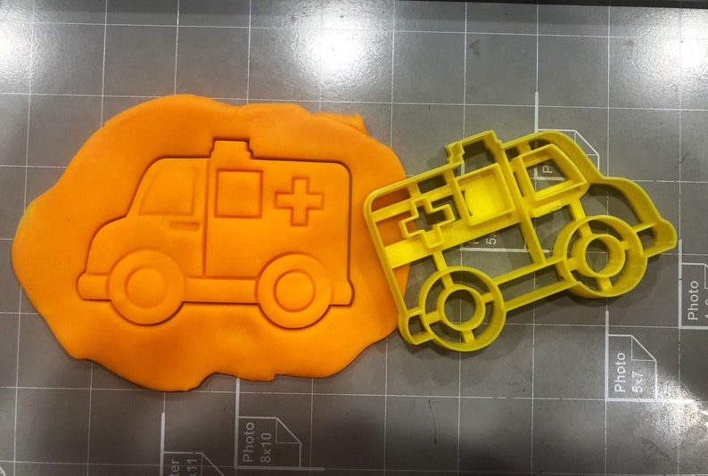 May include: Yellow plastic cookie cutter shaped like an ambulance. The cutter has a red cross on the side and a white rectangular window.