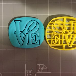 K&ouml;nnte beinhalten: Zwei Ausstecher, einer blau und einer gelb, beide in Form des Wortes "LOVE" in einer fetten, blockartigen Schrift.