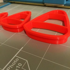 May include: Three red plastic cookie cutters shaped like candy corn. The cutters are on a gray cutting mat with a white "Photo 8x10" label.