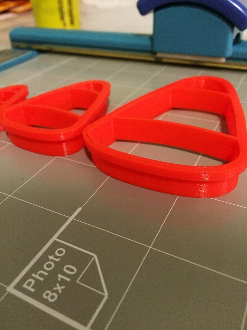 May include: Three red plastic cookie cutters shaped like candy corn. The cutters are on a gray cutting mat with a white "Photo 8x10" label.
