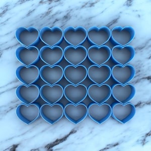 May include: A set of 25 blue heart-shaped cookie cutters arranged in a grid pattern on a white marble surface.