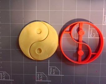 Może przedstawiać: Czerwona plastikowa foremka do ciasteczek w kształcie symbolu yin yang. Foremka do ciasteczek leży na szarej powierzchni z siatką. Obok foremki do ciasteczek leży żółty wykrój ciasta.