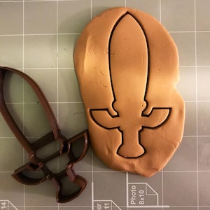 May include: A brown cookie cutter in the shape of a sword with a handle and a cross guard. The cookie cutter is on a brown gridded surface.