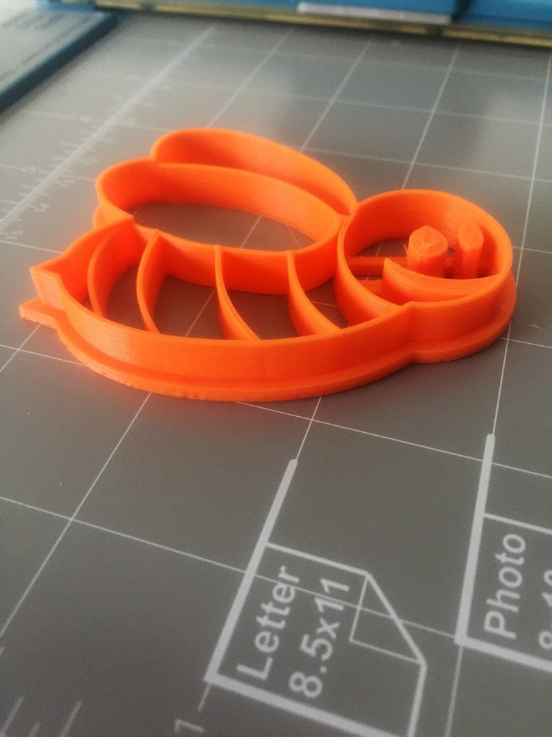 May include: Orange plastic bee-shaped cookie cutter. The cutter has a detailed design with wings and a face.