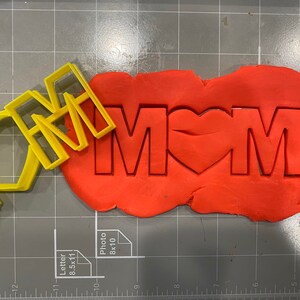 May include: Yellow plastic cookie cutters in the shape of the words "I love Mom" with a heart in place of the "O". The cutters are on top of a red piece of cookie dough.