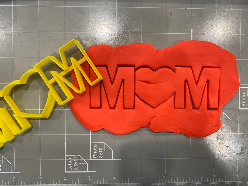May include: Yellow plastic cookie cutters in the shape of the words "I love Mom" with a heart in place of the "O". The cutters are on top of a red piece of cookie dough.