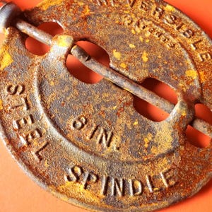 May include: A circular, rusted metal damper plate featuring a central steel spindle. The surface is embossed with the text REVERSIBLE, 6 IN., and STEEL SPINDLE. The object shows significant oxidation and a textured, weathered brown and orange finish.
