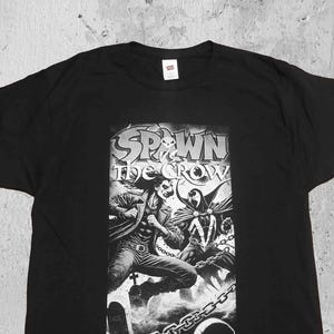 Könnte beinhalten: Schwarzes T-Shirt mit einem weißen Grafikdruck der Figuren Spawn und The Crow. Der Text "Spawn the Crow" ist über den Figuren gedruckt.