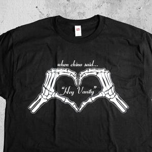 May include: Black t-shirt featuring a white skeleton hand heart design with the text "when chino said..." and "Hey Vanity". The shirt is a crew neck style.