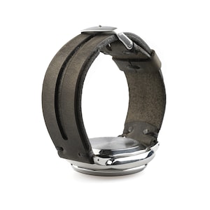 May include: A wristwatch with a silver case and a wide, dark grey leather strap. The strap has a double-layer design and a silver buckle. The watch is seen from the side, highlighting the strap's texture and the watch face.