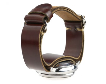 Puede incluir: Una correa de reloj de cuero marrón con una esfera de reloj de acero inoxidable. La correa es gruesa y se envuelve alrededor de la muñeca. La esfera del reloj es redonda y tiene un acabado plateado. La correa está cosida con hilo de color claro.