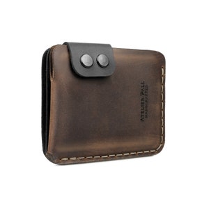 May include: A brown leather wallet with a black leather flap secured by two black metal snaps. The wallet has white stitching around the edges and the text "ATELIER PALL HANDCRAFTED" is printed vertically on the front.