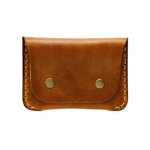 May include: A small, rectangular, brown leather wallet with a flap closure secured by two brass-colored snap buttons. The edges are stitched with a contrasting light-colored thread, and the leather has a textured appearance.
