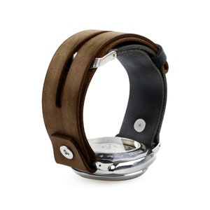 May include: A brown leather watch band with a stainless steel watch face. The band has a double-strap design and a black leather backing. The watch face is silver and has a detailed dial.