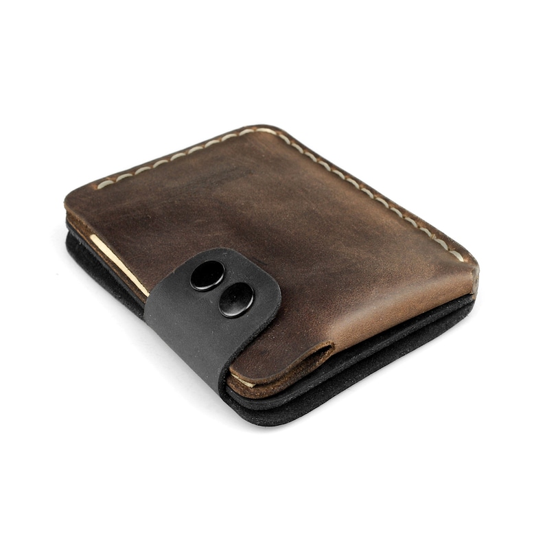 May include: A brown leather wallet with a black strap secured by two black snaps. The wallet has a stitched border with cream-colored thread. The wallet appears to be made of multiple layers of leather and fabric.