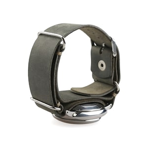 Op de afbeelding: Een grijze leren horlogeband met een roestvrijstalen horlogekast. De band heeft een breed bandontwerp met zilverkleurige metalen hardware. De horlogekast is rond en heeft een zilveren afwerking. De band is ontworpen om om de pols te wikkelen.