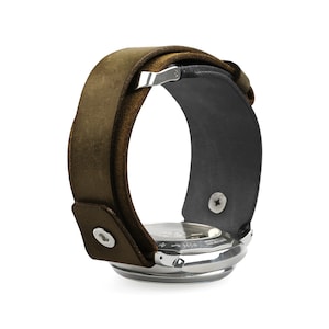 Puede incluir: Un reloj plateado con una ancha correa de cuero marrón. La esfera del reloj es visible, y la correa se envuelve, asegurada con un cierre metálico y un cierre a presión. El reloj tiene un diseño clásico y robusto.