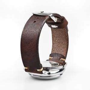 Può includere: Un orologio da polso con cinturino in pelle marrone e quadrante argentato. Il cinturino è cucito con filo colorato chiaro e ha una fibbia argentata. Il quadrante è rotondo e ha una lunetta argentata.