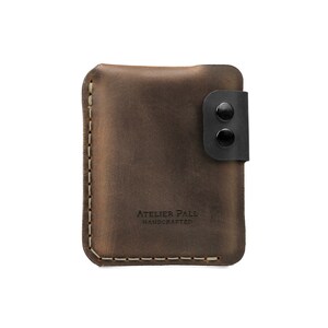 May include: A brown leather wallet with a black snap closure. The wallet has a stitched border and the text "ATELIER PALL HANDCRAFTED" is embossed on the front. The wallet is rectangular and appears to be made of high-quality leather.