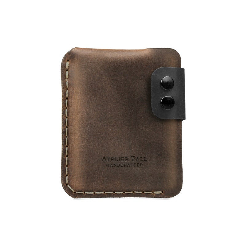 May include: A brown leather wallet with a black snap closure. The wallet has a stitched border and the text "ATELIER PALL HANDCRAFTED" is embossed on the front. The wallet is rectangular and appears to be made of high-quality leather.